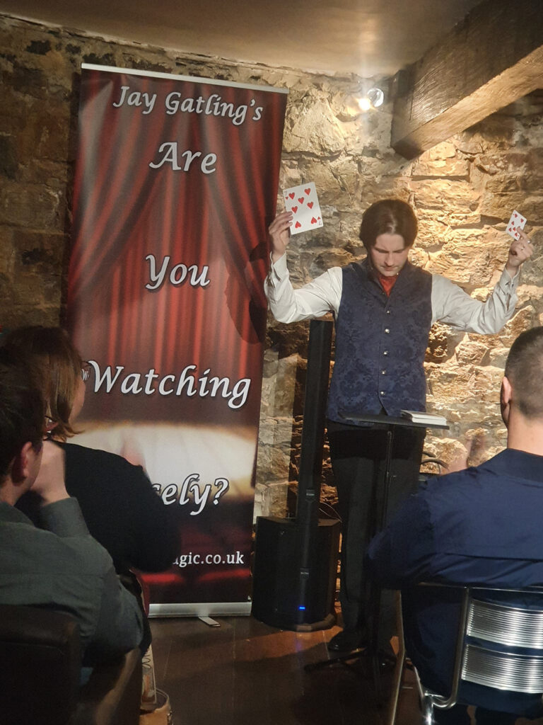 Magician Jay Gatling holds a jumbo playing card in one hand, and a regular card in the other hand. He is standing in front of a banner with the words "Are You Watching Closely?" There are stone walls and wooden beams in the background.