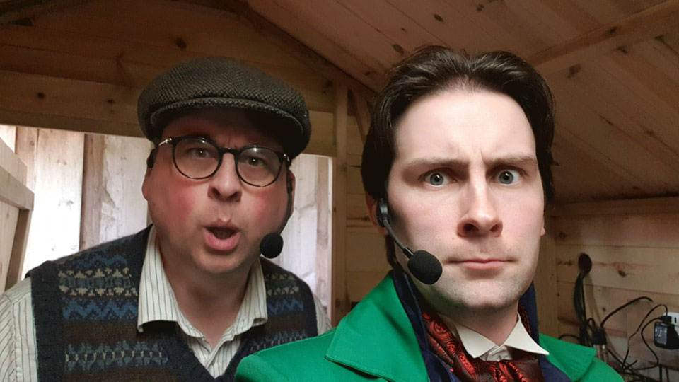A man with glasses and a flat cap stands behind a man with a bright green jacket. They are both wearing headset microphones, and standing in what appears to be a shed.