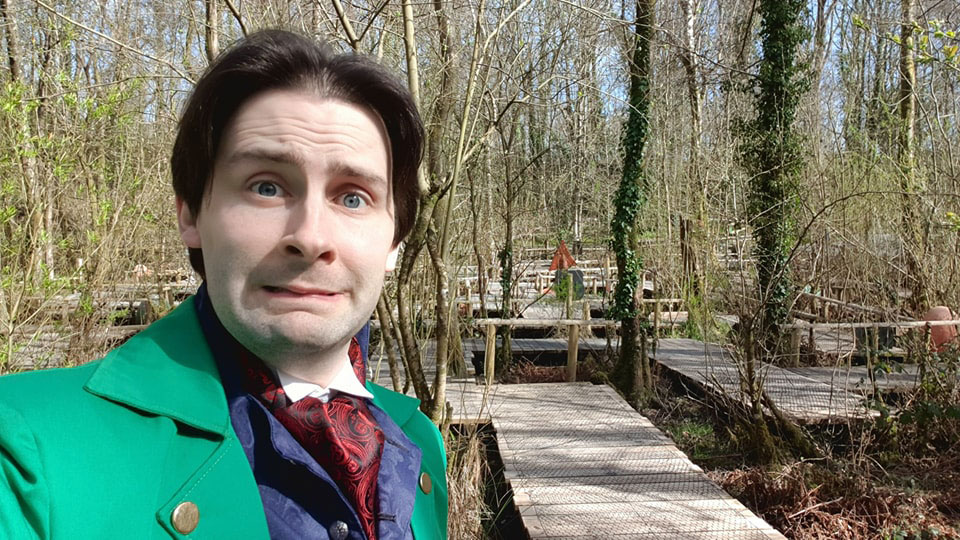 A man with dark hair and a bright green jacket is pulling a face at the camera. Behind him is a board walk maze set among numerous trees.